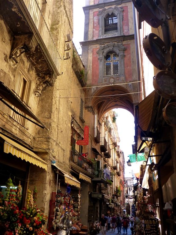 a narrow alleyway in a european city, with tall buildings on either side. an archway connects two buildings above the street. people are walking down the street, and there are shops and stalls along the sides. flowers are displayed in front of a shop on the left. tambourines are hanging on the right. balconies extend from the buildings above. the scene captures the lively atmosphere of a bustling urban environment.
