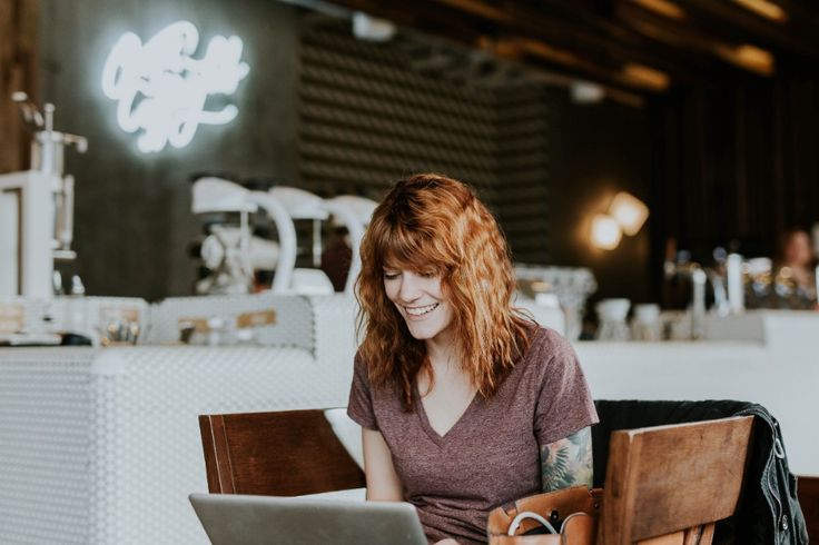 a young woman with red hair and a tattoo on her arm sits at a table in a cafe, smiling and looking at a laptop. a neon sign and coffee machine are visible in the background.