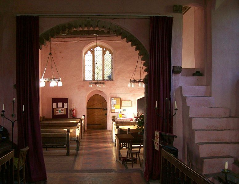the interior of a church is depicted, featuring rows of wooden pews on either side of a tiled aisle. at the far end of the aisle is a wooden door and a window. two chandeliers hang from the ceiling. the entrance to the church is framed by dark red curtains and an ornate archway. to the right of the entrance, there is a staircase built into the wall. the walls are painted a muted pink. tables and chairs are visible in the background, suggesting a multipurpose space. the overall impression is of a historic and somewhat dimly lit religious setting.