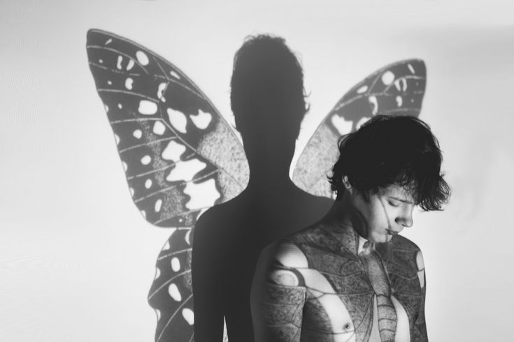 a man with a butterfly shadow projected on him.  the shadow of the butterfly covers the man's back and wings  the butterfly's wings are black and white, and the man is  facing the camera.  he's looking down and his hair is covering his eyes. the man's torso is covered in black and white body paint that appears to be tattooed on his skin.  a creative fine art black and white photo.