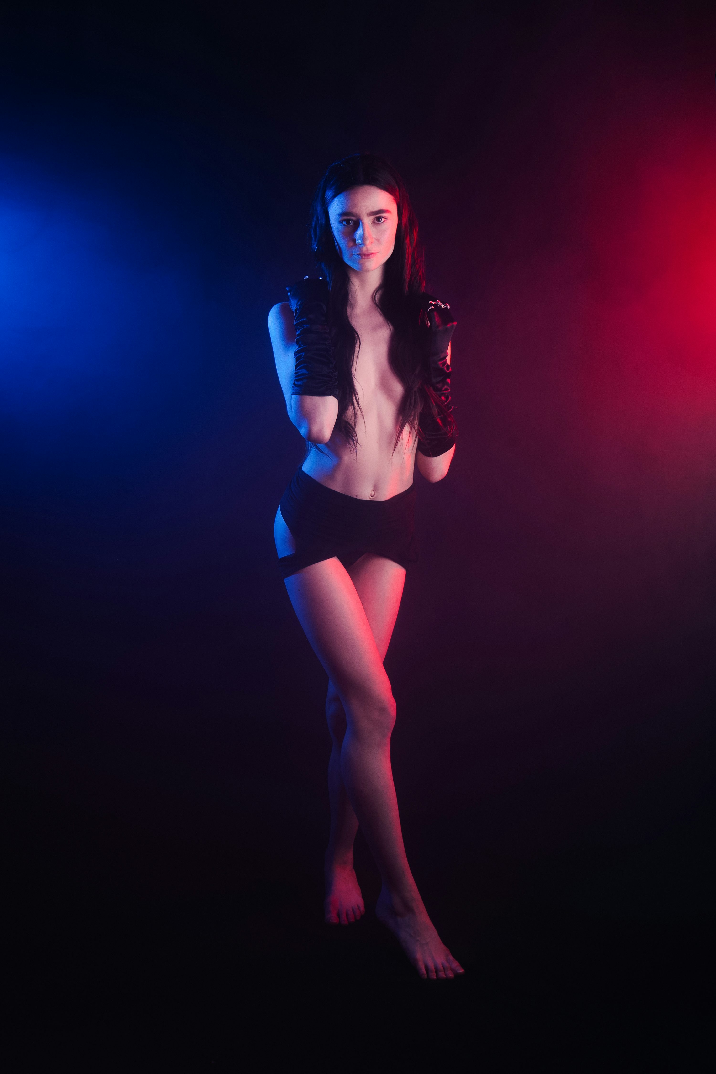 a captivating portrait of a topless woman in a short black skirt and black gloves. she stands confidently against a dark background illuminated with red and blue lighting, creating a sultry and mysterious atmosphere. her body is slightly angled, showcasing her long legs and graceful posture. the lighting enhances the overall mood and creates interesting shadows, adding depth to the image.