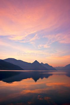 the image is of a mountain range silhouetted against a sunset sky. the mountains are reflected in a lake in the foreground. the colors of the sky are pink, orange, and purple. the water in the lake is calm and reflective. the overall mood of the image is peaceful and serene.