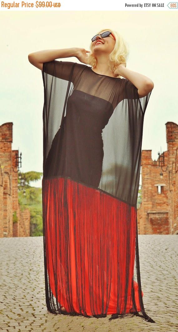 a young woman wearing a red and black dress, poses outdoors. the dress is a sheer black fabric with a red fringe at the bottom. the woman has long blonde hair and is wearing sunglasses. she is standing with her arms raised above her head, in front of brick buildings.