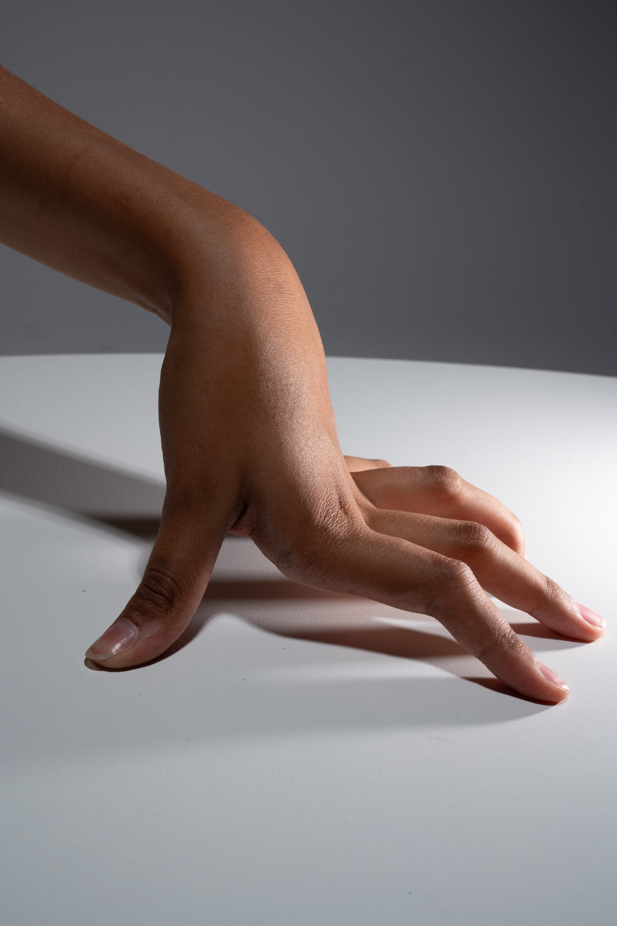 a closeup image of a hand with dark skin tone, gently touching a white table surface. the hand is illuminated with studio lighting, creating a contrast between the warm skin tones and the cool white of the table.