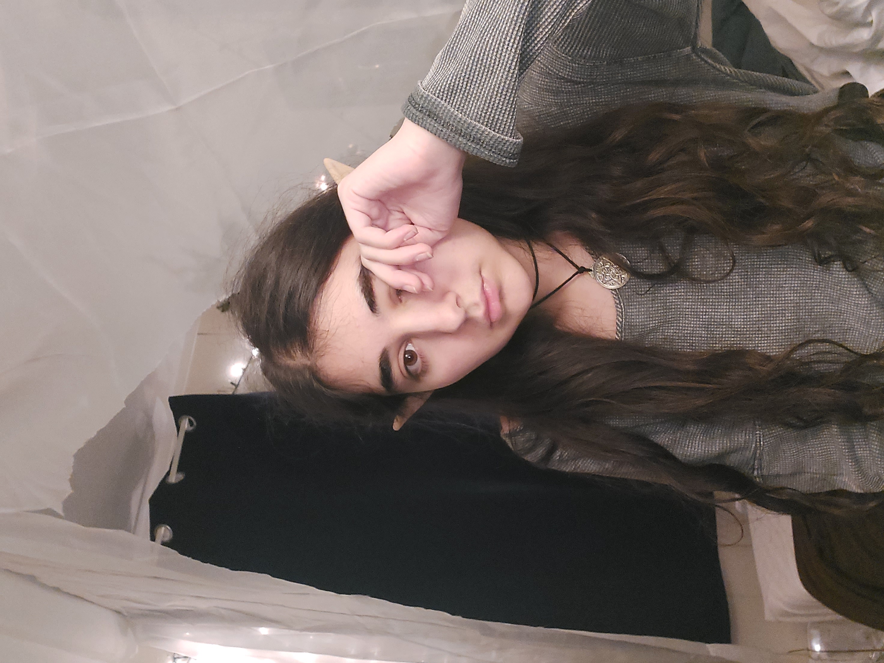 the image shows a teenage girl with long, dark brown, wavy hair wearing elf ears.  she's dressed in a gray, longsleeved sweater and a simple necklace. she appears to be indoors in a bedroomlike setting, partially obscured by a white fabric drape and dark curtains. she has a somewhat sad or pensive expression and is wiping her eye with her hand.  the overall mood is slightly moody and intimate. the setting appears cozy, and there's a soft, warm light from fairy lights visible in the background. the photo seems like a selfportrait or cosplay image.