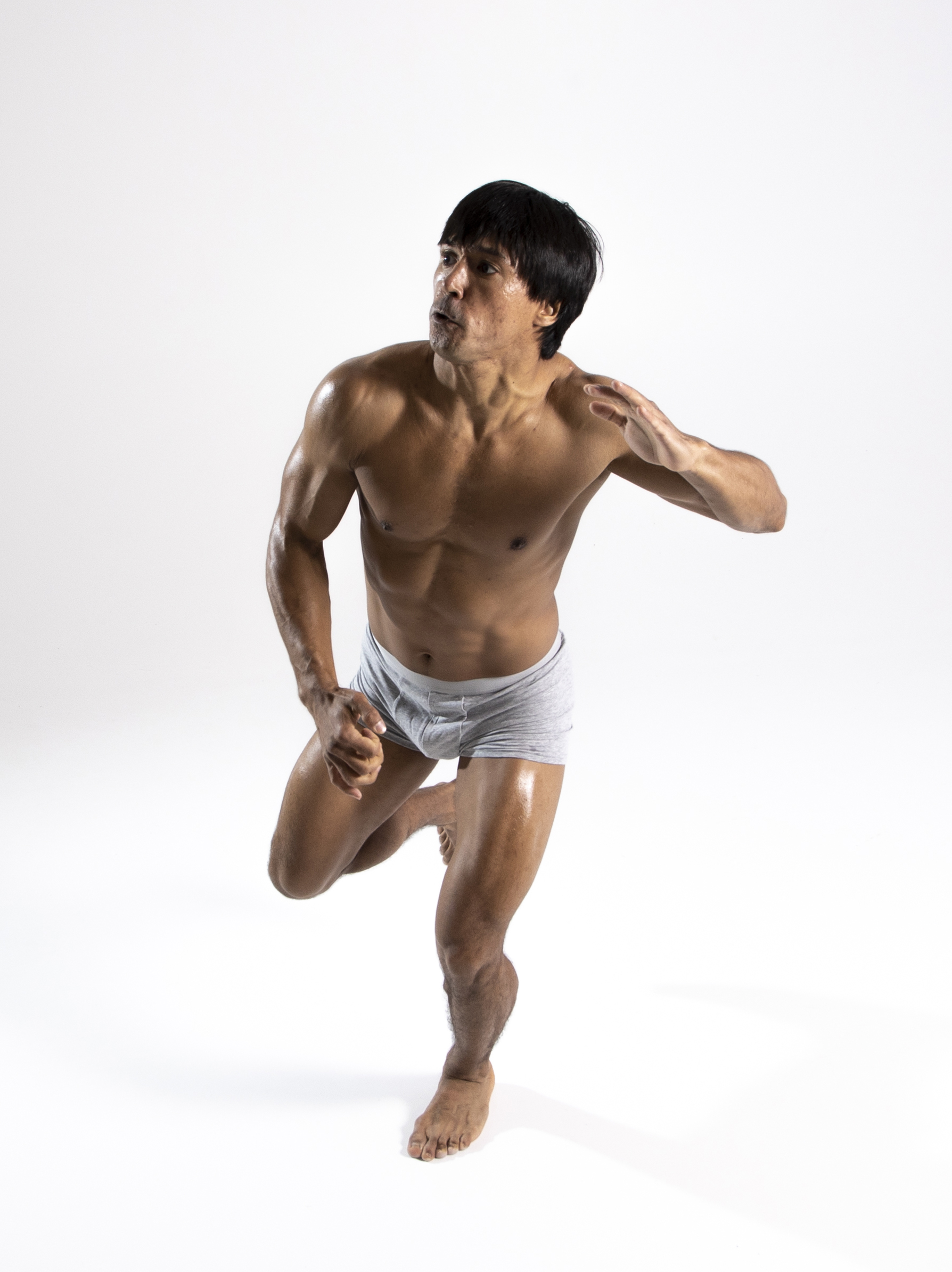 the image shows a muscular, shirtless man wearing underwear, in a running pose against a white background. he appears athletic with a toned physique and oiled skin. he is barefoot, and the pose conveys motion and energy.