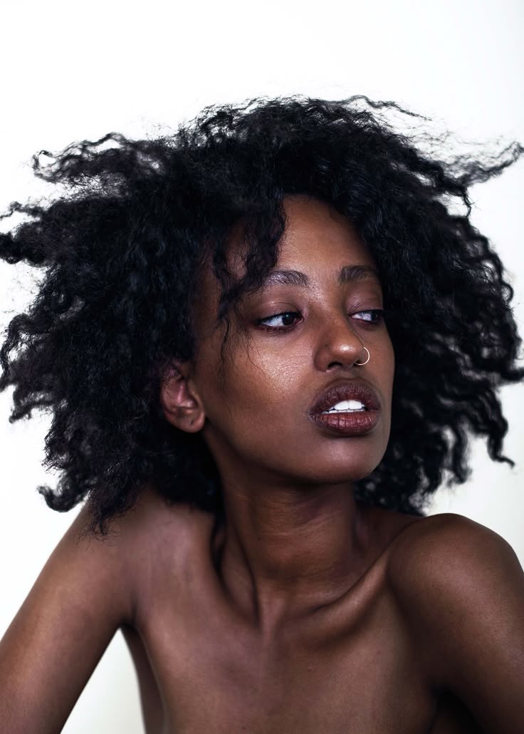 a striking portrait of a woman with dark, voluminous afrotextured hair and a nose ring, looking off to the side against a white background. her expression is thoughtful, capturing a sense of depth and allure. her shoulder is bare.