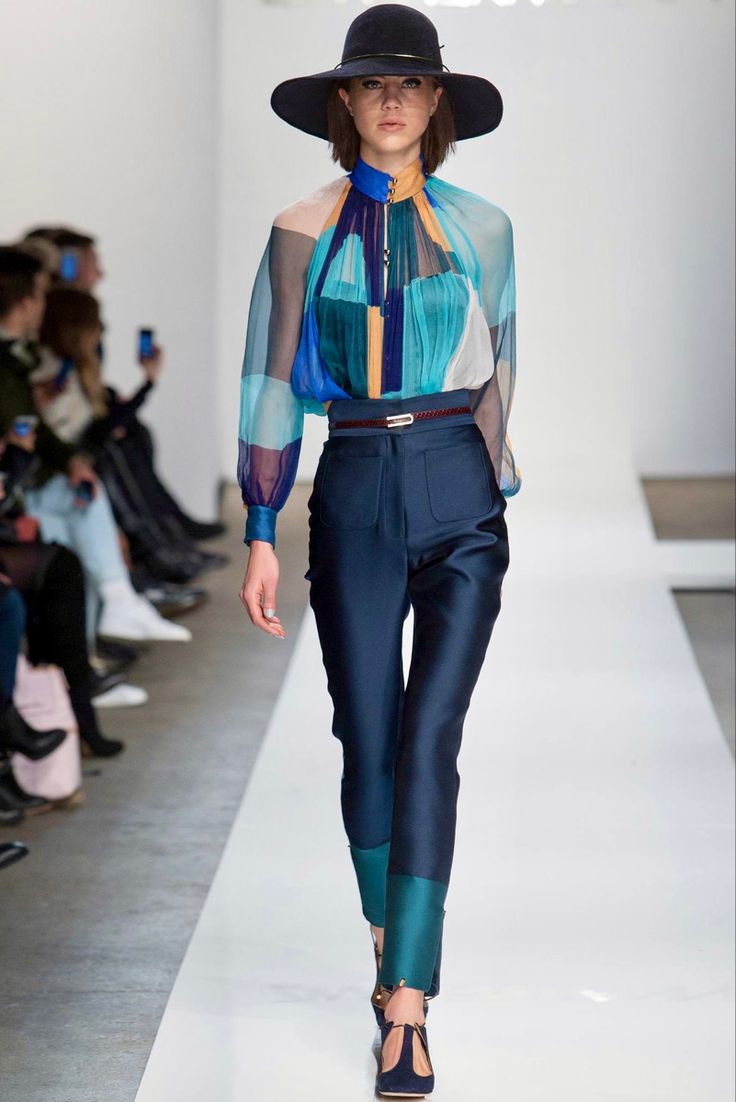 a female model walks the runway wearing a black hat, blue and turquoise blouse, dark blue pants with green trim, and black shoes. she is walking towards the camera. the background is white and there are other models in the background.