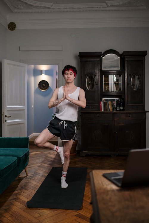 a young man with red hair and a headband is practicing yoga in his living room. he is standing on one leg, balancing himself, in a tree pose.  he's wearing white socks with red stripes, black shorts, and a white tank top.  a laptop is on a nearby table, suggesting he might be following an online workout class. the room is furnished with a dark wooden cabinet and a dark green sofa. the hardwood floors and antique furniture give the setting a classic, slightly vintage feel. the overall mood is one of peace and tranquility.