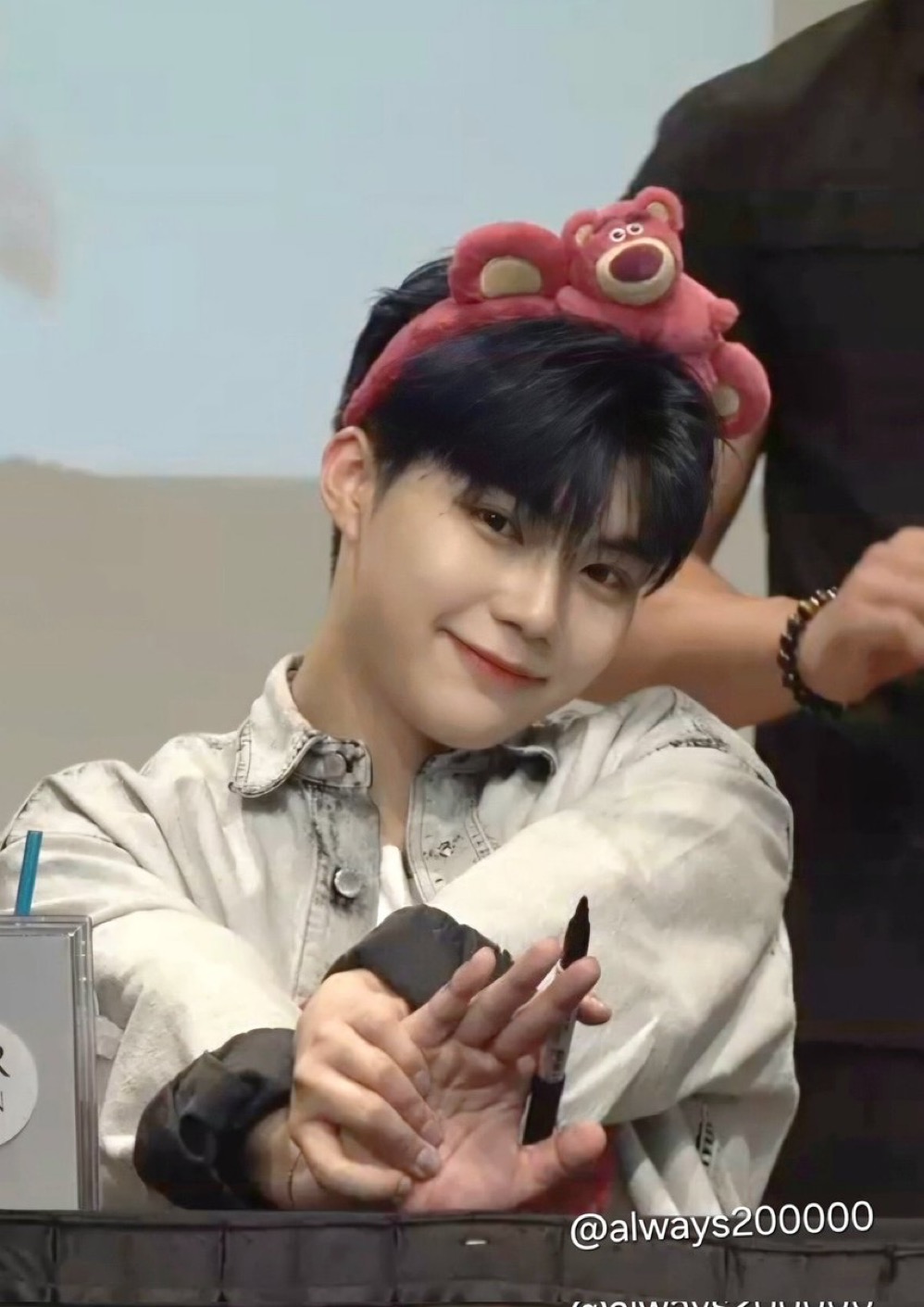 a closeup shot of a young man with dark hair and a headband sitting at a table and signing autographs. he is wearing a light gray denim jacket over a white shirt. the environment and his pose suggest that he is a kpop idol at a fansign event.