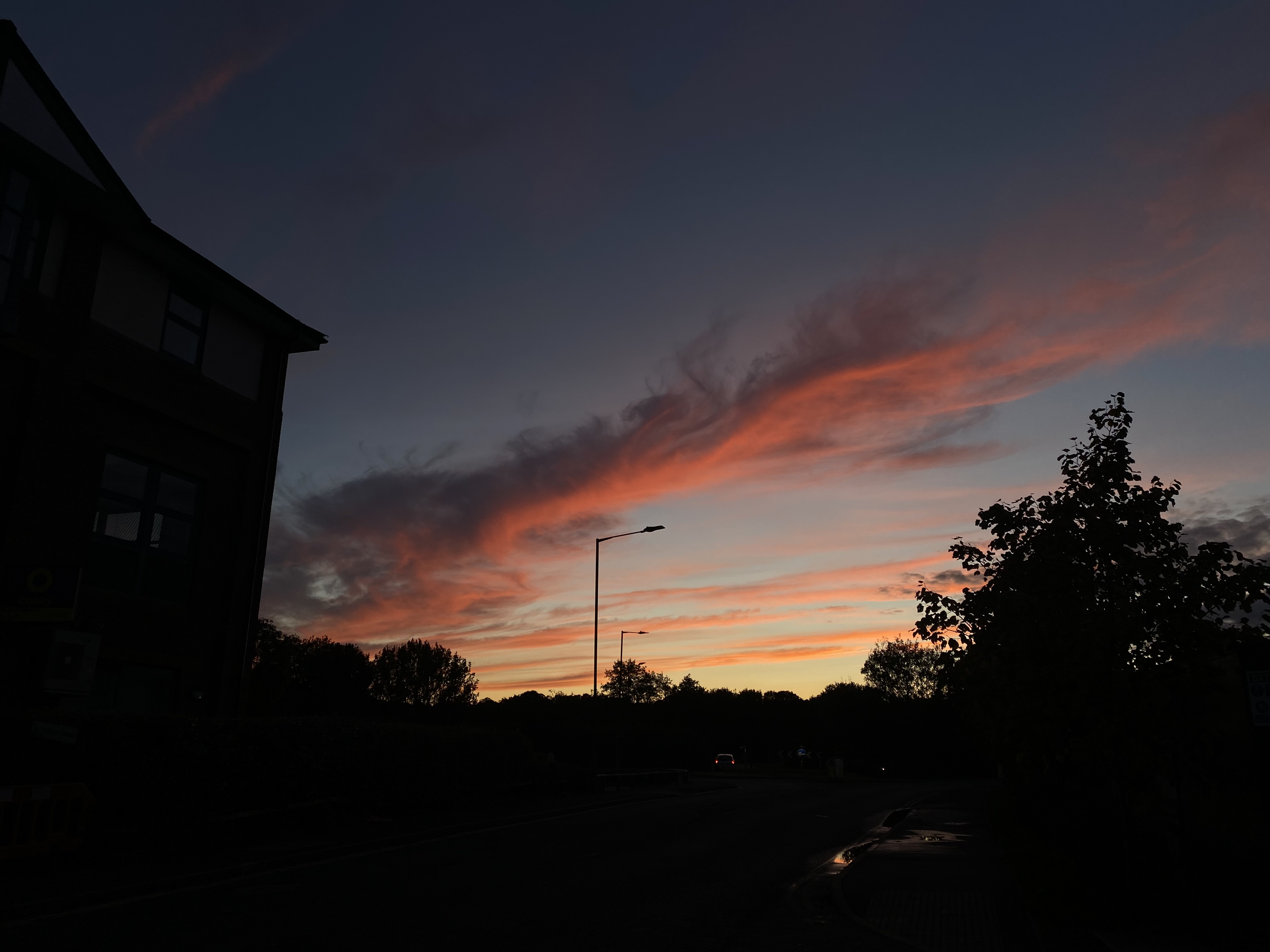 a dramatic sunset sky with vibrant orange, pink, and red hues fills the frame. silhouetted against the sky are buildings, trees, and streetlights. the scene is captured from a street level perspective, with the road and pavement in the foreground. the overall atmosphere is peaceful and serene, creating a beautiful landscape of nature and urban elements.