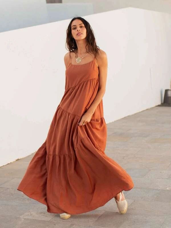 a woman is posing outdoors wearing a long, terracottacolored linen maxi dress with spaghetti straps. she has a necklace and sandals. she is standing in front of a white wall.