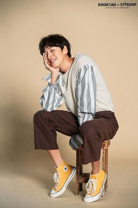 a korean man wearing a striped shirt, brown pants and yellow sneakers is sitting on a wooden chair smiling at the camera. he looks stylish and casual.