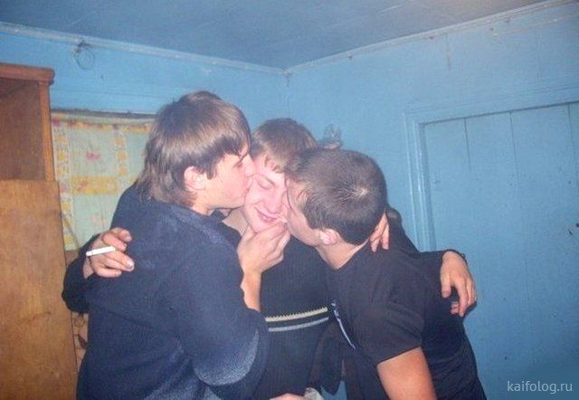 three young men stand indoors in front of a blue wall. they are close together and appear to be hugging and kissing. the man in the middle is the focus of the photo, and the two men on either side are leaning in and touching him on the face. the three are smiling and look happy.