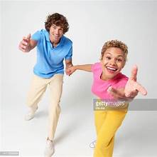 a young interracial couple is captured in middance move against a clean white background. the woman, with short blonde hair and wearing a vibrant pink and yellow outfit, smiles brightly as she reaches towards the camera. the man, with brown curly hair and a blue and white ensemble, playfully extends his hand towards hers. the image evokes a sense of joy, energy, and connection.