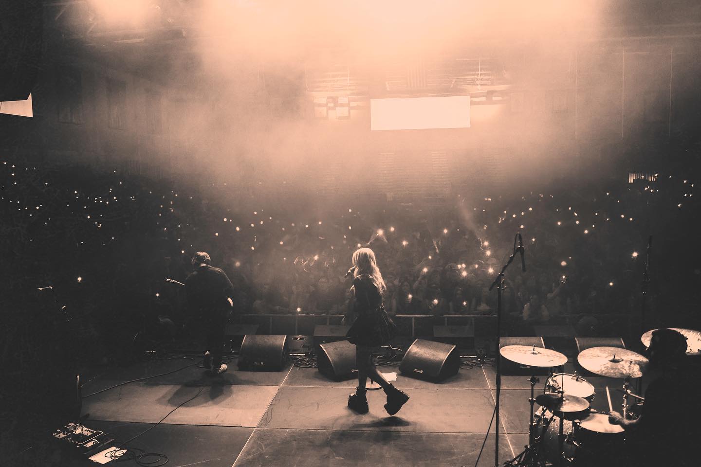 a grayscale shot of a concert performance. a female singer in a short dress and platform boots is singing into a microphone in the center of the stage. to her left, a guitarist is playing, and to her right, a drummer is behind a drum set. the stage is illuminated with bright lights, and there is fog creating a hazy atmosphere. the audience in the background is holding up phones with their flashlights on, creating a sea of tiny lights.