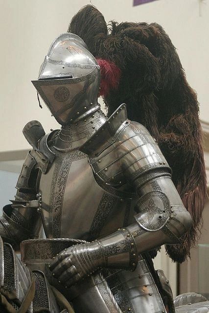 a closeup view of a knight's suit of armor, likely on display in a museum.  the armor is made of polished metal plates, intricately crafted and engraved.  the knight's helmet is particularly ornate, featuring a raised visor and a plume of dark feathers. the lighting is soft and highlights the metallic sheen and details of the armor.
