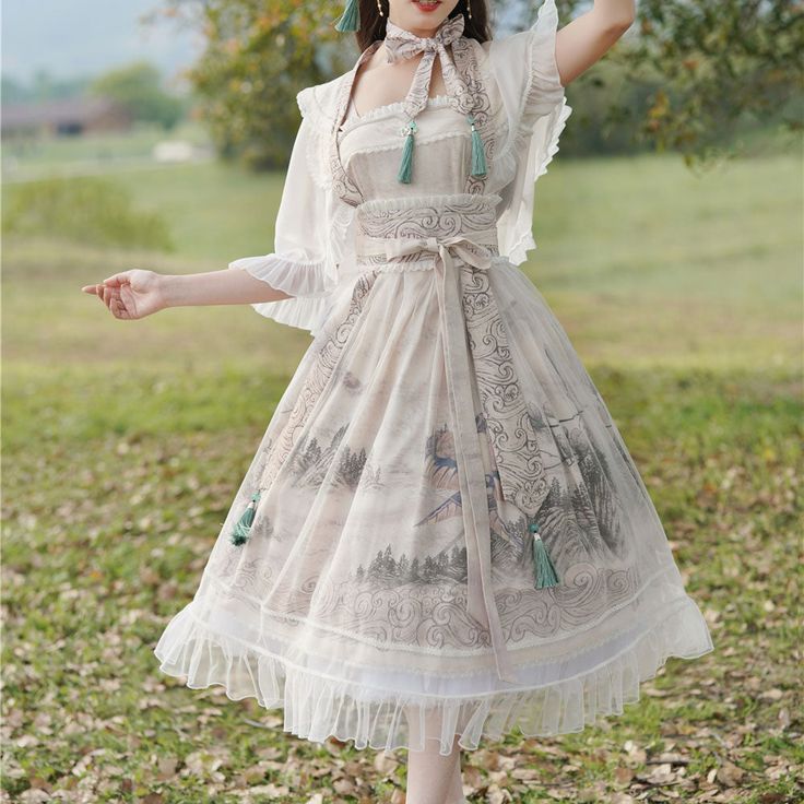a woman wearing a flowing white dress with a nature scene printed on the skirt. the dress is layered and includes a white top, an overlayer with delicate straps, and a belt accented with tassels. the photo is taken outdoors in natural light and shows the woman from the knees up as she looks off to the side.