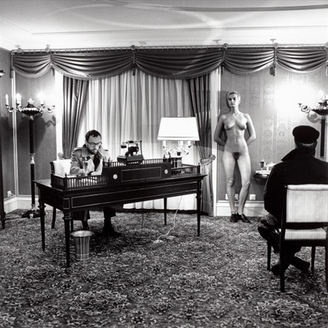 the black and white photograph depicts a scene in what appears to be a 1970sera hotel room.  a man in a suit sits at a dark wood desk, seemingly working or taking a phone call.  a naked woman stands silently against a wall behind him. a third individual, dressed in dark clothing, is seated in a chair observing the scene.  the overall atmosphere is quiet and somewhat tense. the image has a stark, almost cinematic quality, suggesting a performance or staged event, rather than a candid shot. the contrasting figures and their contrasting postures create visual tension. the setting, complete with curtains and a table lamp, adds to the somewhat formal and controlled atmosphere.