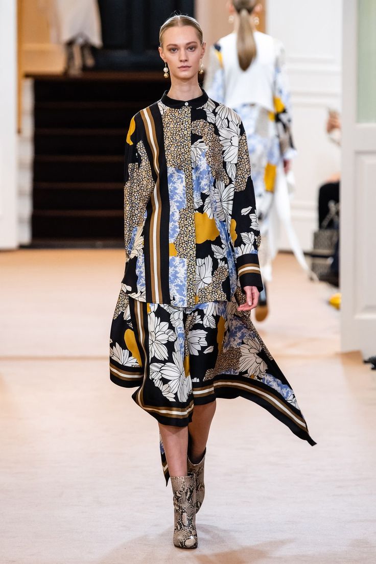 a model walks down a runway wearing a stylish black and gold floral patterned dress and snake print ankle boots. the dress has a unique design with different fabric panels and patterns, creating a patchwork effect.  the model has her hair pulled back in a sleek style, and minimal makeup. the background shows a blurred out view of other models and the interior of a fashion show venue. the overall mood is sophisticated and elegant.
