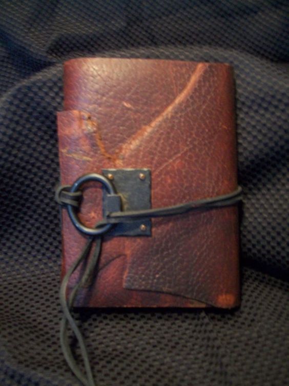 this is an image of a handmade, leatherbound journal. it's crafted from distressed brown leather with visible natural markings. a black leather cord wraps around the journal, securing it with a metal loop and a square metal plate.  the journal rests on a textured, dark background.