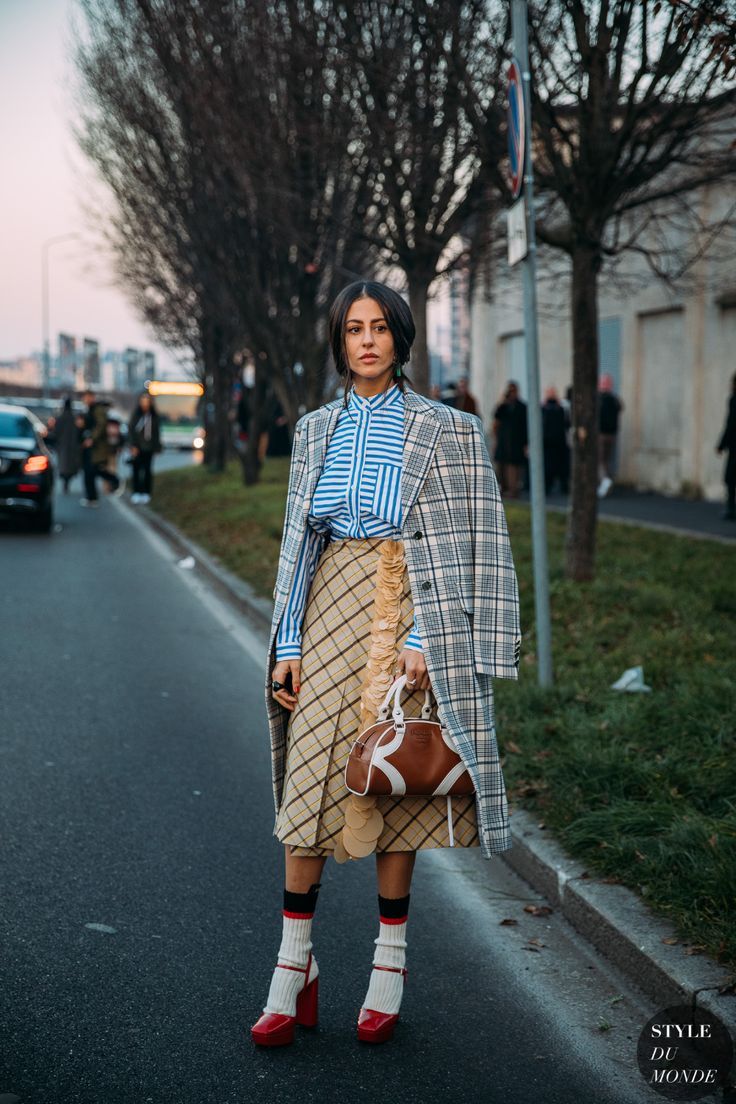 a stylish woman strolls down a milan street, her outfit a blend of textures and patterns.  she wears a grey plaid overcoat over a blue and white striped shirt and a beige pleated midi skirt.  her accessories include red chunky heeled shoes with cream socks, and a brown and white handbag.  the backdrop is a typical european city scene with trees and buildings. the overall aesthetic is chic and elegant, capturing the essence of street style fashion.