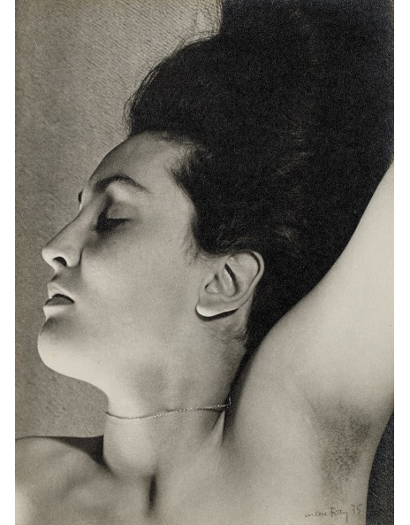 the image is a black and white closeup portrait of a woman reclining. her hair is styled in a high, dark updo. her eyes are closed, and her expression is serene. she is nude from the shoulders up, and a thin necklace is visible around her neck. the lighting is dramatic, focusing on her face and upper body. the photograph has a timeless quality and an artistic style reminiscent of 1930s surrealism. the signature of man ray is visible in the bottom right corner.  the overall feel is elegant, sensual, and somewhat mysterious.