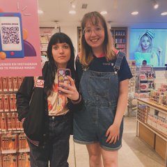 two young women pose for a selfie in a brightly lit store aisle. they are both stylishly dressed, one in a denim jumpsuit and the other in a black bomber jacket and graphic tee. the image captures a sense of friendship and the experience of shopping in a modern retail environment. the aisles are filled with various products and promotional materials, including a poster with a qr code.
