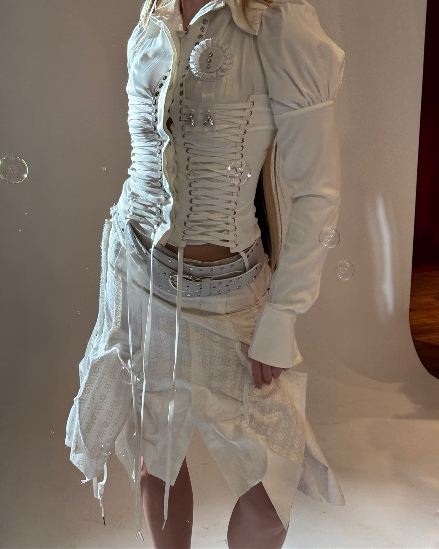 a woman is modeling a white fashion outfit. she is wearing a white blouse, a white corset, and a skirt. bubbles are floating in the air around her.