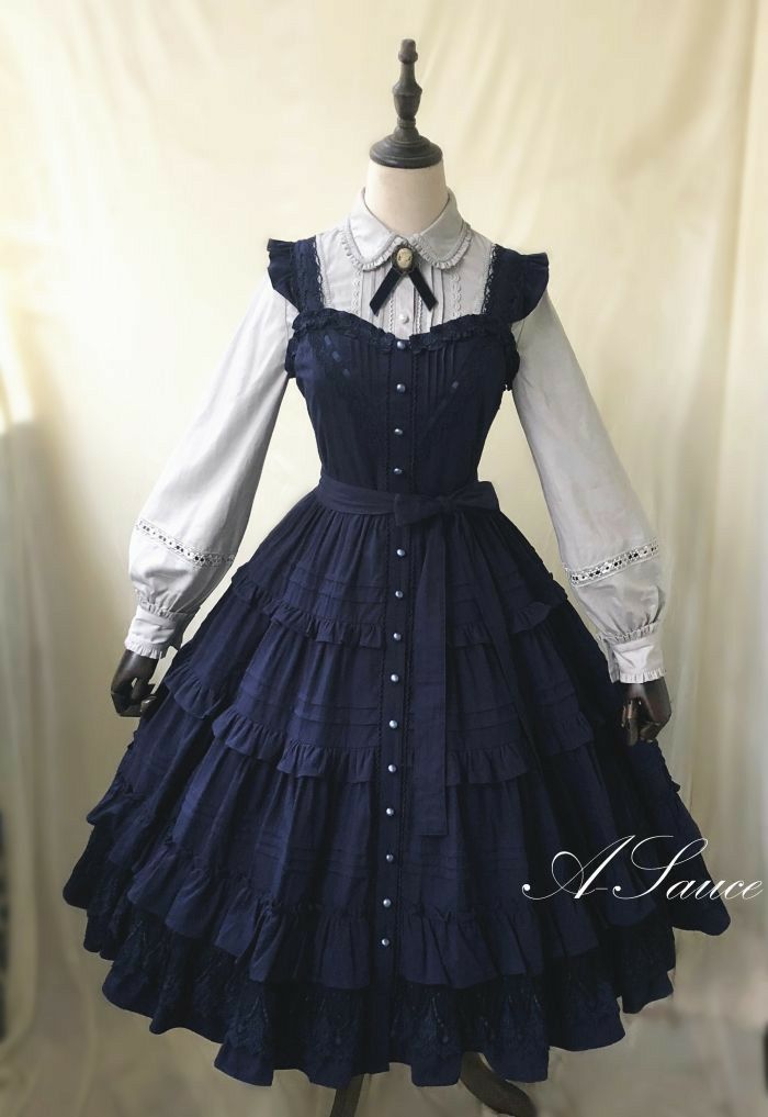 this is a picture of a navy blue lolita fashion dress on a mannequin. the dress has a white blouse with a peter pan collar worn underneath. the jumper skirt has a high waist, buttons down the front, and a tiered skirt with ruffles. the dress is accessorized with a bow at the collar.