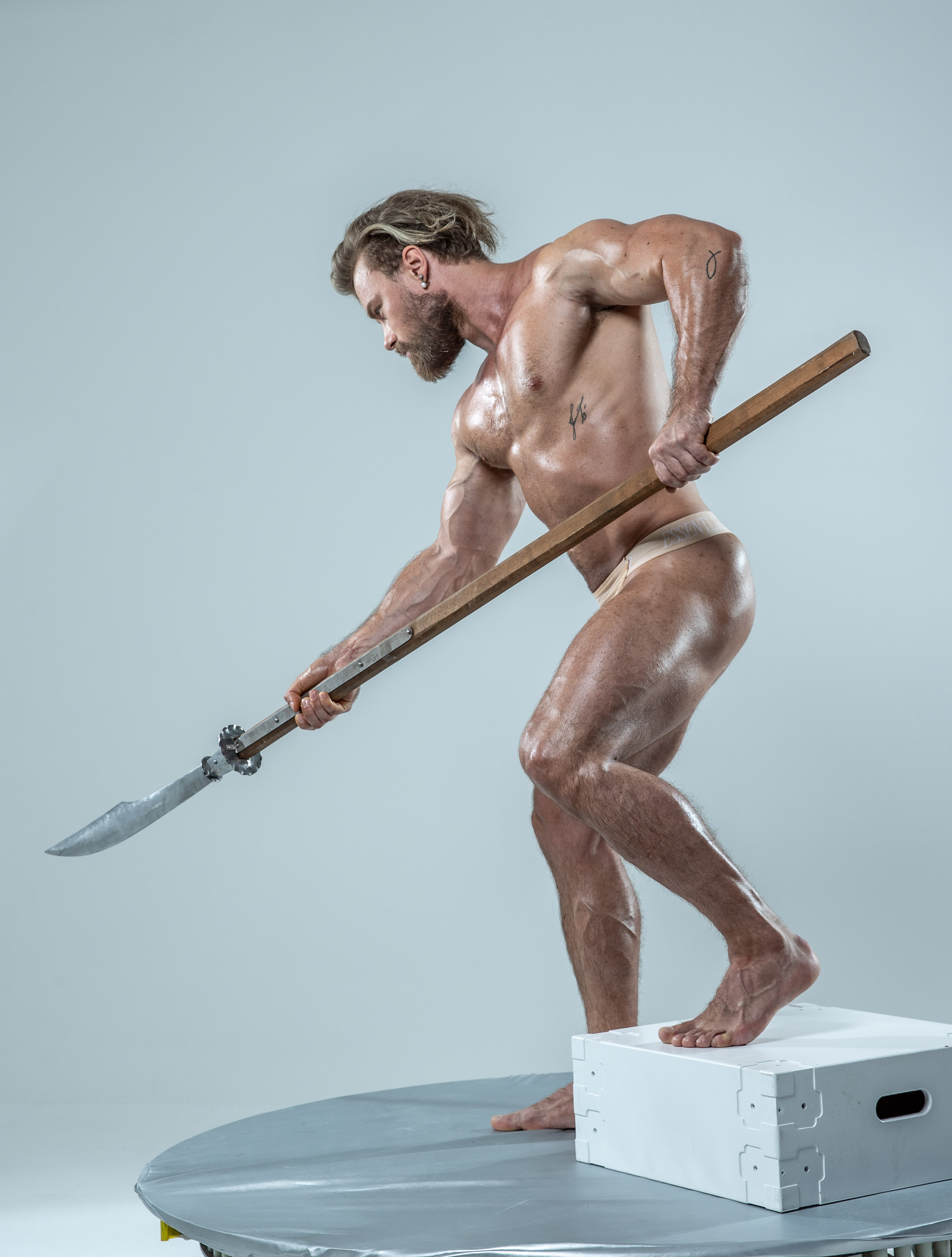 a muscular, oiled man with long blond hair and a beard holds a spear aggressively in a studio setting. he stands on a white platform with a circular base, positioned against a light gray backdrop. the low camera angle emphasizes his powerful stance and the dynamic tension of the pose.  his physique and the spear evoke classical imagery of warriors and gladiators.  the studio lighting is soft and even, highlighting the definition of his muscles.