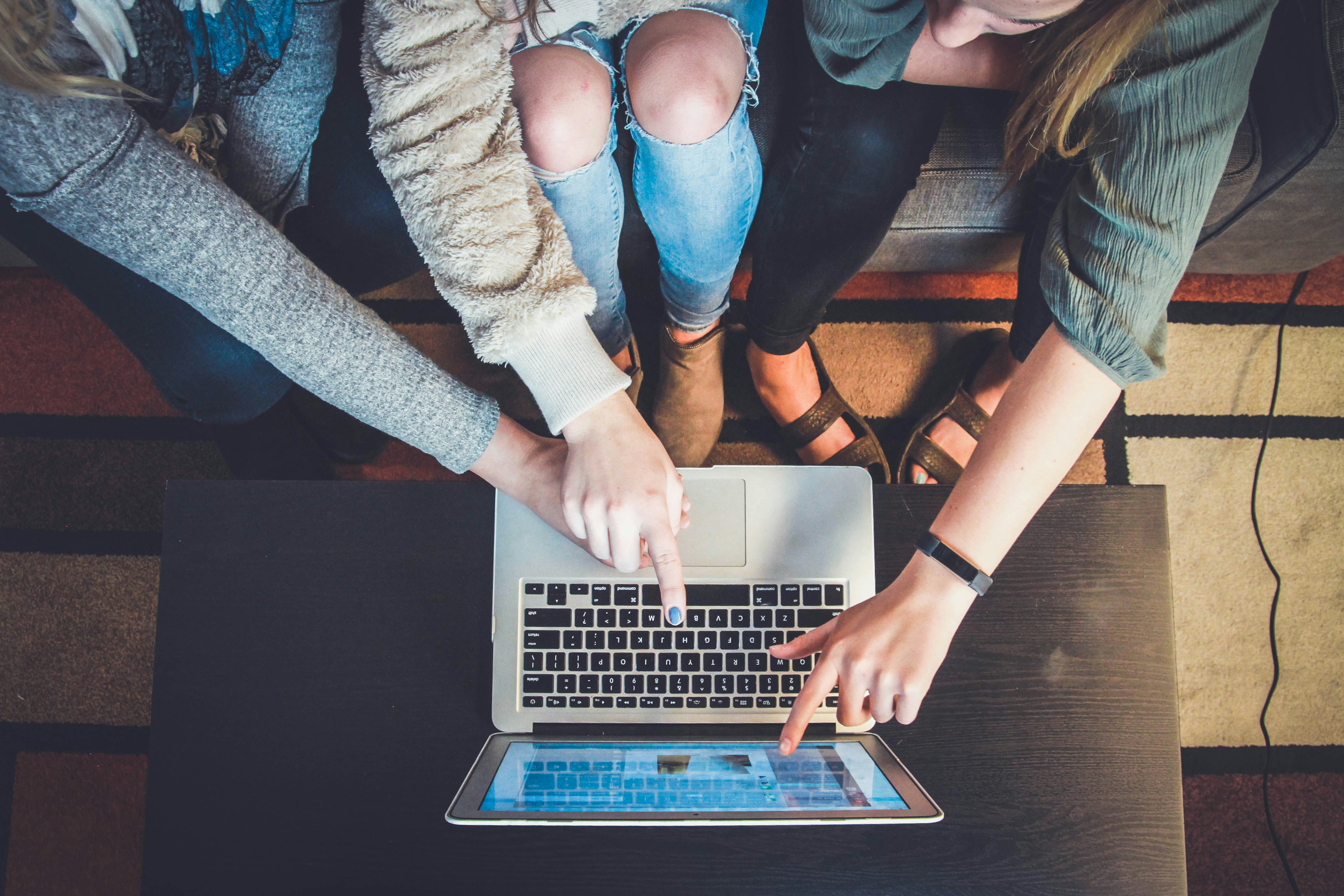 the image shows three women sitting on a couch and gathered around a laptop computer. they are looking at the screen and pointing at it with their fingers, suggesting they are working on something together. the image is taken from a topdown perspective and features a warm and inviting atmosphere.