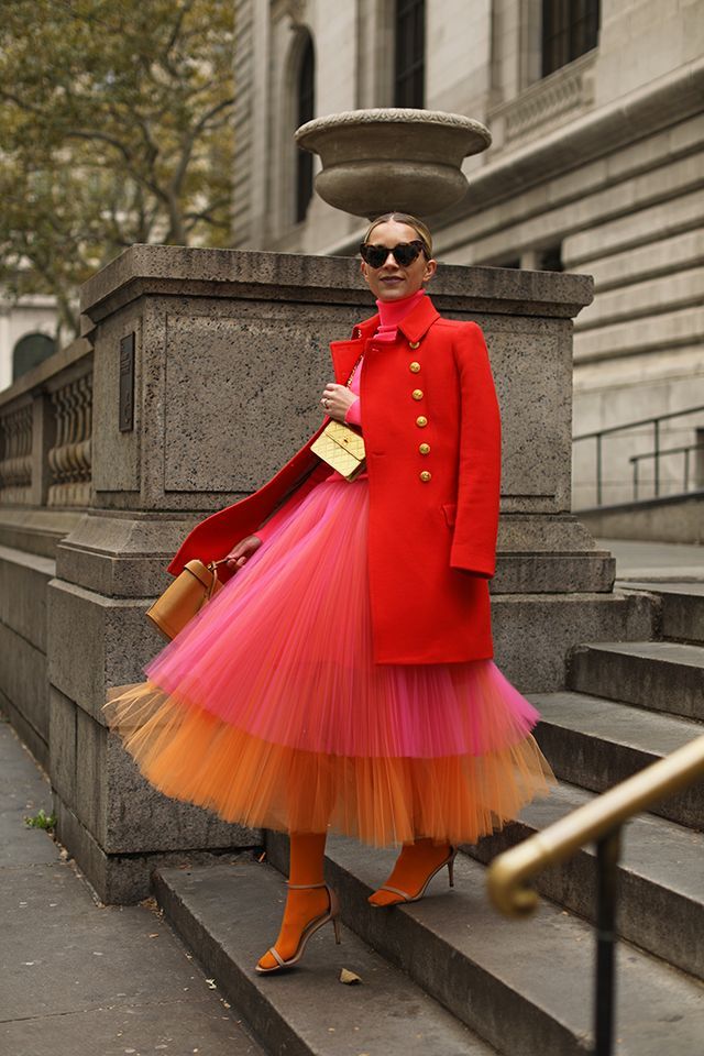 a woman in a red coat, pink and orange tulle skirt, gold purse and sunglasses is walking down stairs in the city. she is wearing heels and looks stylish and trendy.