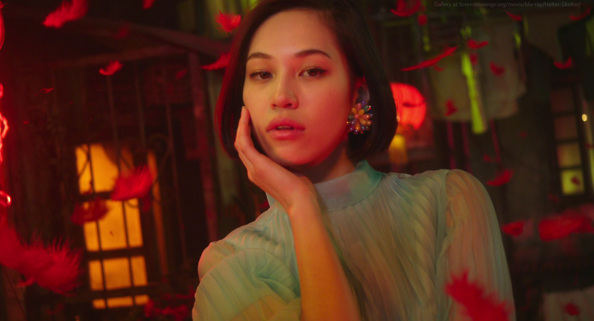 a young woman with short dark hair is looking directly at the camera. she is wearing a blue and green dress with a soft glow. she has red feathers flying around her.  her  skin is glowing and she is wearing  delicate earrings.  the image is soft and elegant.