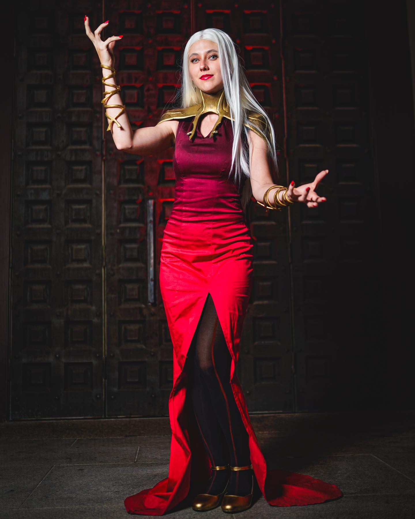 a striking photograph of a woman cosplaying a character, possibly a sorceress or magical being. she is wearing a floorlength red dress with a maroon bodice, detailed gold jewelry including armbands and a choker, and black heels. her long white hair contrasts sharply with the rich colors of her attire. the background is a dark, imposing doorway, enhancing the mystical and dramatic atmosphere. her pose suggests power and confidence, her hands elegantly raised as if performing a magical gesture. the overall mood is mysterious and captivating, hinting at a fantastical narrative. the image's lighting and composition contribute to its artistic and cinematic quality.