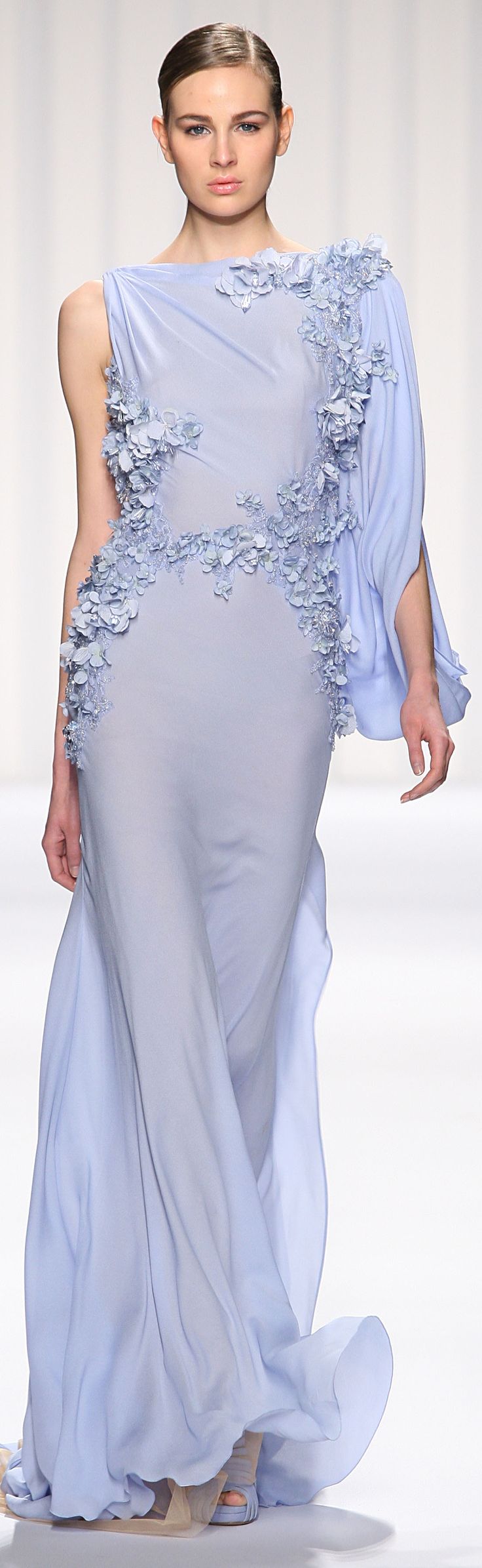 a female model walks the runway in a light blue gown with a floral embellishment, a flowing train, and white platform shoes. the dress is very elegant and the model is posing with one hand on her hip. the background is a white wall.