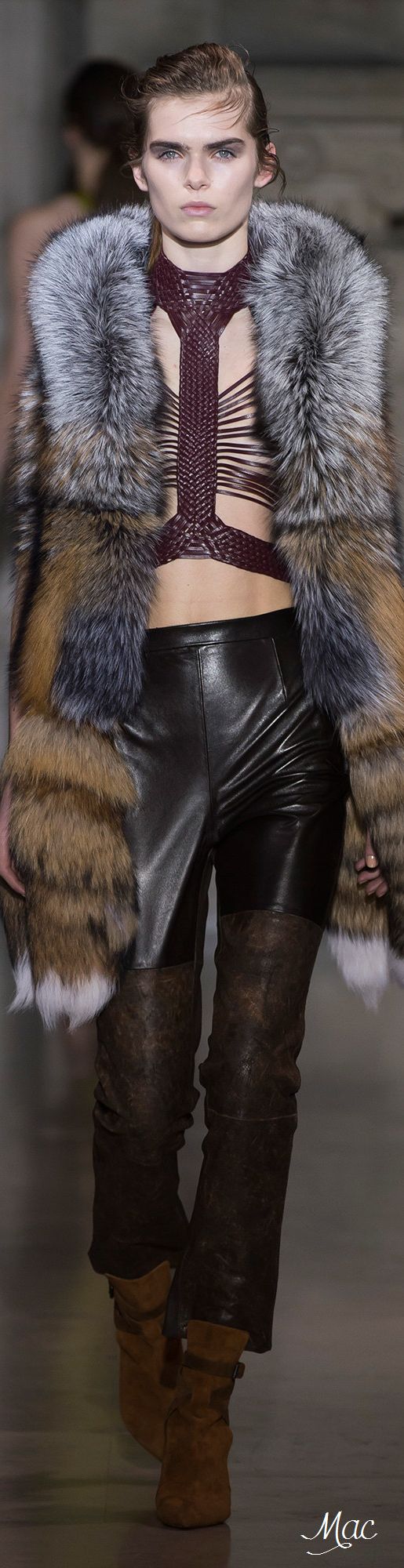 a woman in a fur coat and leather pants poses on the runway. she is wearing a dark fur coat with a silver lining and black leather pants. she is also wearing brown boots and has her hair pulled back in a ponytail. she is looking at the camera with a confident expression. the backdrop of the runway is a blurred out grey space.