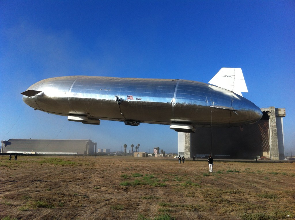 a silver blimp is hovering in the air over a grass field with a hangar building behind it. the blimp is tethered to the ground. the hangar is open and the blimp is partially inside the hangar. there are people walking on the field below the blimp. the sky is blue with a few white clouds. there are trees and a small forest in the background. the blimp has a us flag on the side.