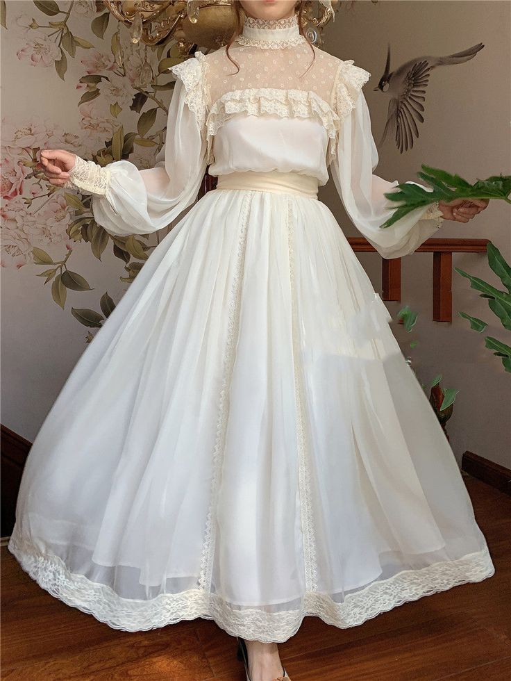 a woman wearing a long white dress with lace detailing. the dress has a high neck, long sleeves, and a flowing skirt. the woman is standing in a room with a floral wallpaper and wooden stairs. the lighting is soft and natural, creating a romantic and dreamy atmosphere.