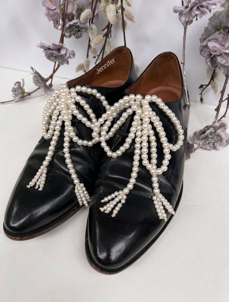 a closeup shot showcases a pair of black leather oxford shoes adorned with delicate pearl embellishments.  the pearls are arranged in cascading strands forming tassels that hang down the front of each shoe. the shoes are positioned on a white surface, and a sprig of silverygray dried flowers is artfully arranged in the background, adding a touch of elegance to the scene. the inside of one shoe is visible, showing the name "jennifer" written inside. the overall image is one of sophisticated style and meticulous detail.