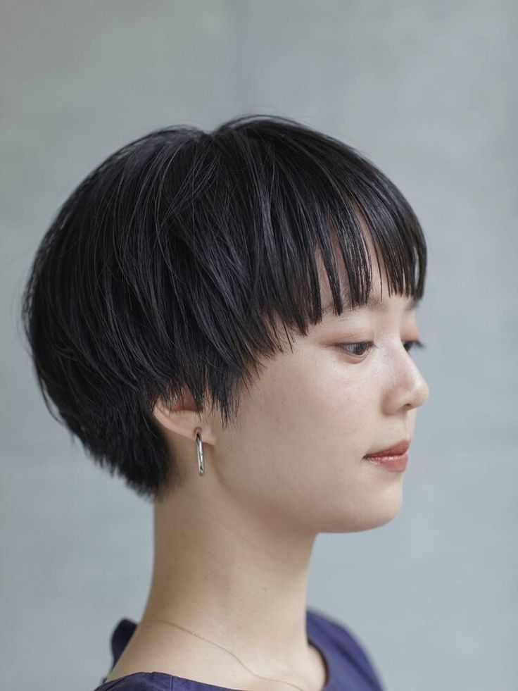a closeup profile shot of a young asian woman with a short black pixie haircut, wearing a silver hoop earring and a black top against a gray background. she is looking slightly away from the camera and has a slightly solemn expression.
