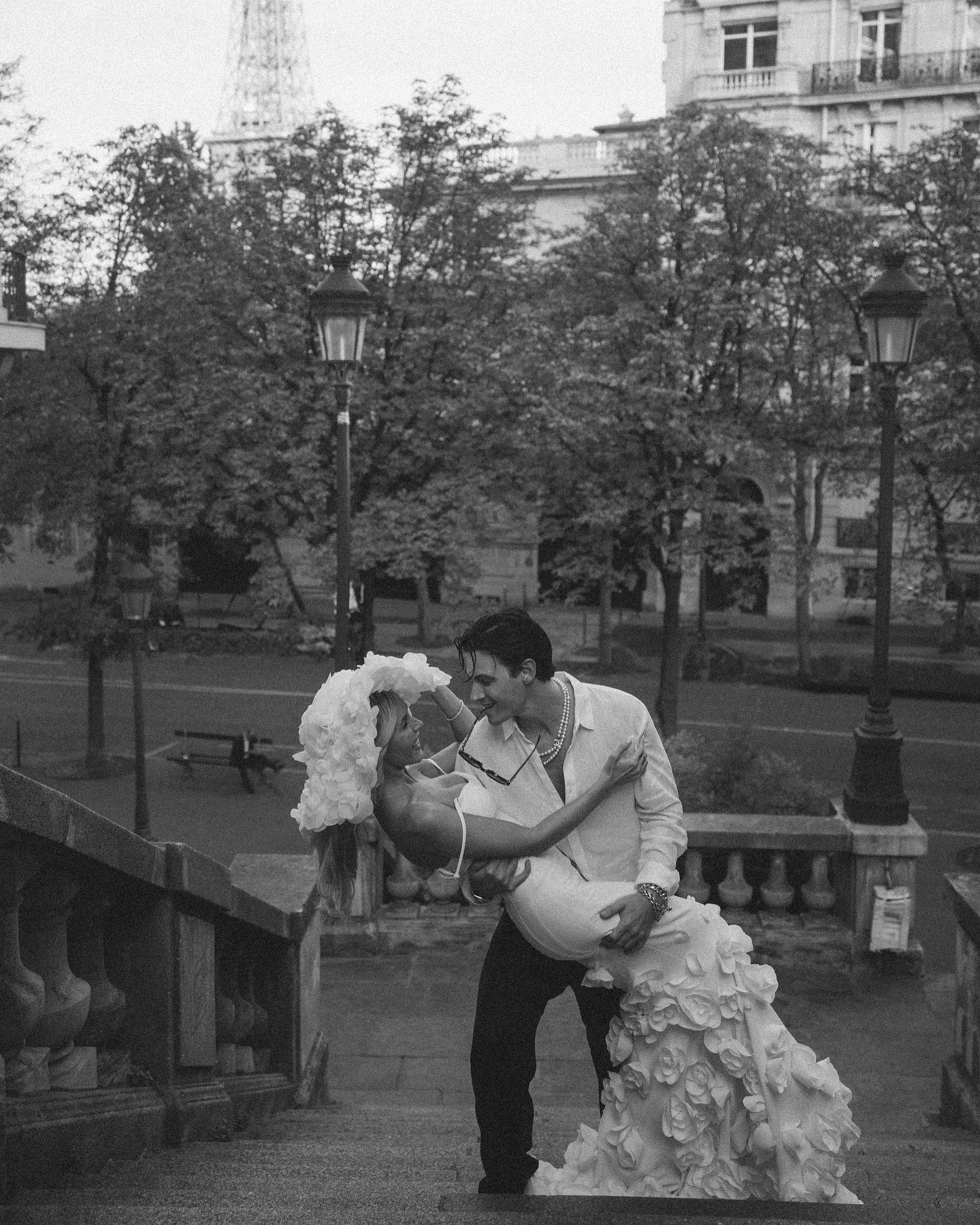 a black and white photo of a couple in love, kissing in the street. the man is wearing a suit and the woman is wearing a white dress. they are standing in front of a building with the eiffel tower in the background. the photo is taken in paris, france.