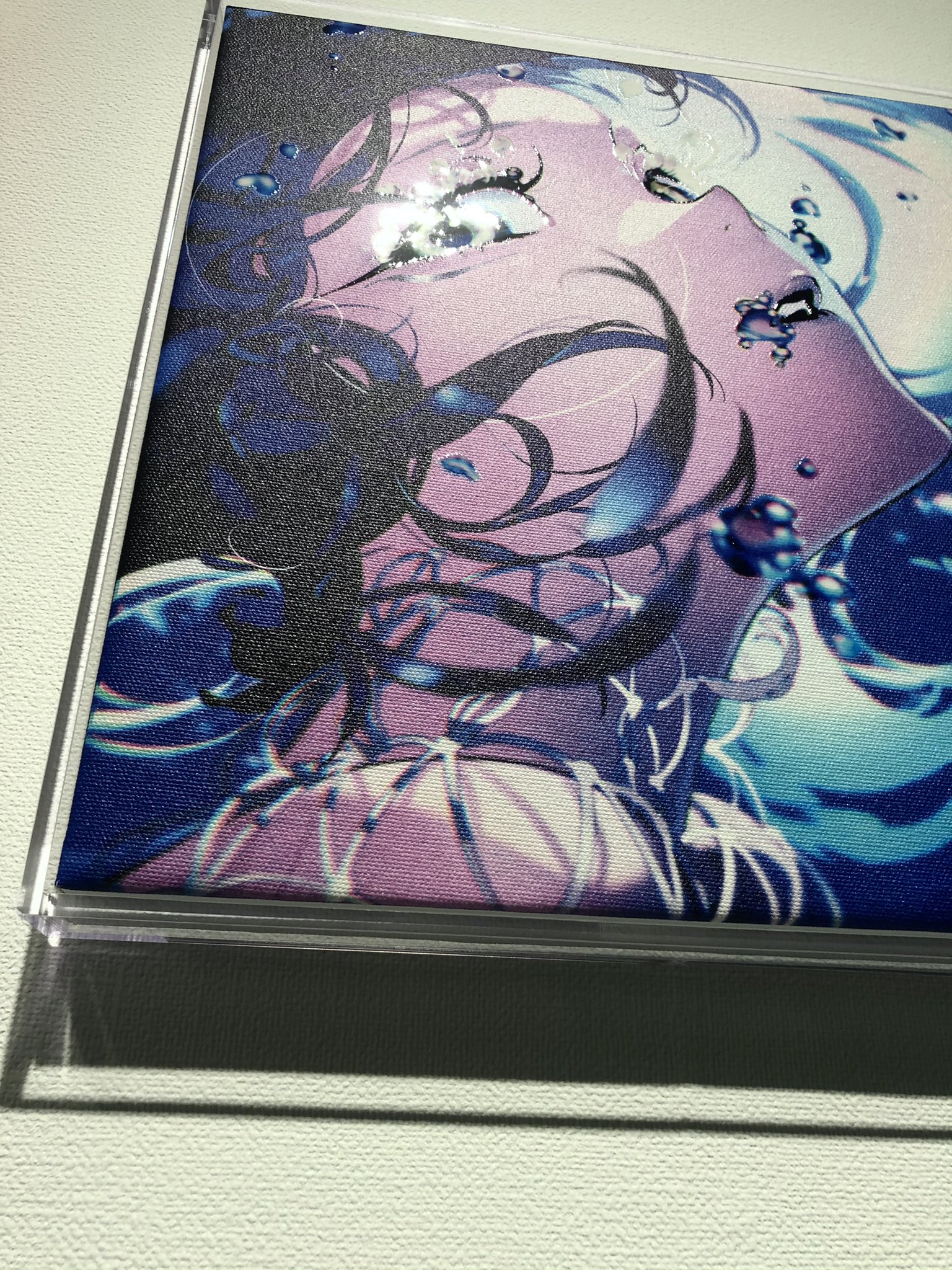 the image shows a closeup of a framed anime illustration featuring a woman with long dark hair submerged in water. the artwork is mounted on a wall and illuminated with indoor lighting. the color palette is dominated by shades of blue and purple.
