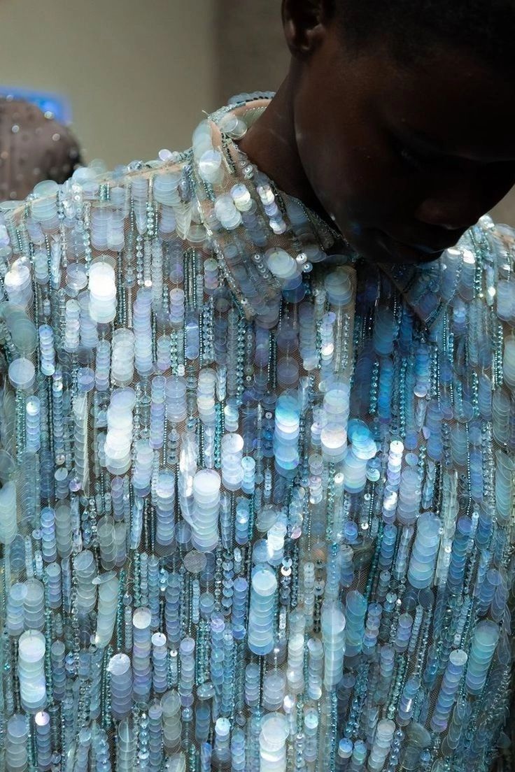 a closeup shot of a woman wearing a sparkling blue and silver sequined dress. the sequins are arranged in a pattern of vertical lines, creating a shimmering effect. the dress is a high fashion piece, and the model is wearing it in a way that makes it look glamorous and elegant. the lighting is soft and diffused, making the sequins sparkle even more.