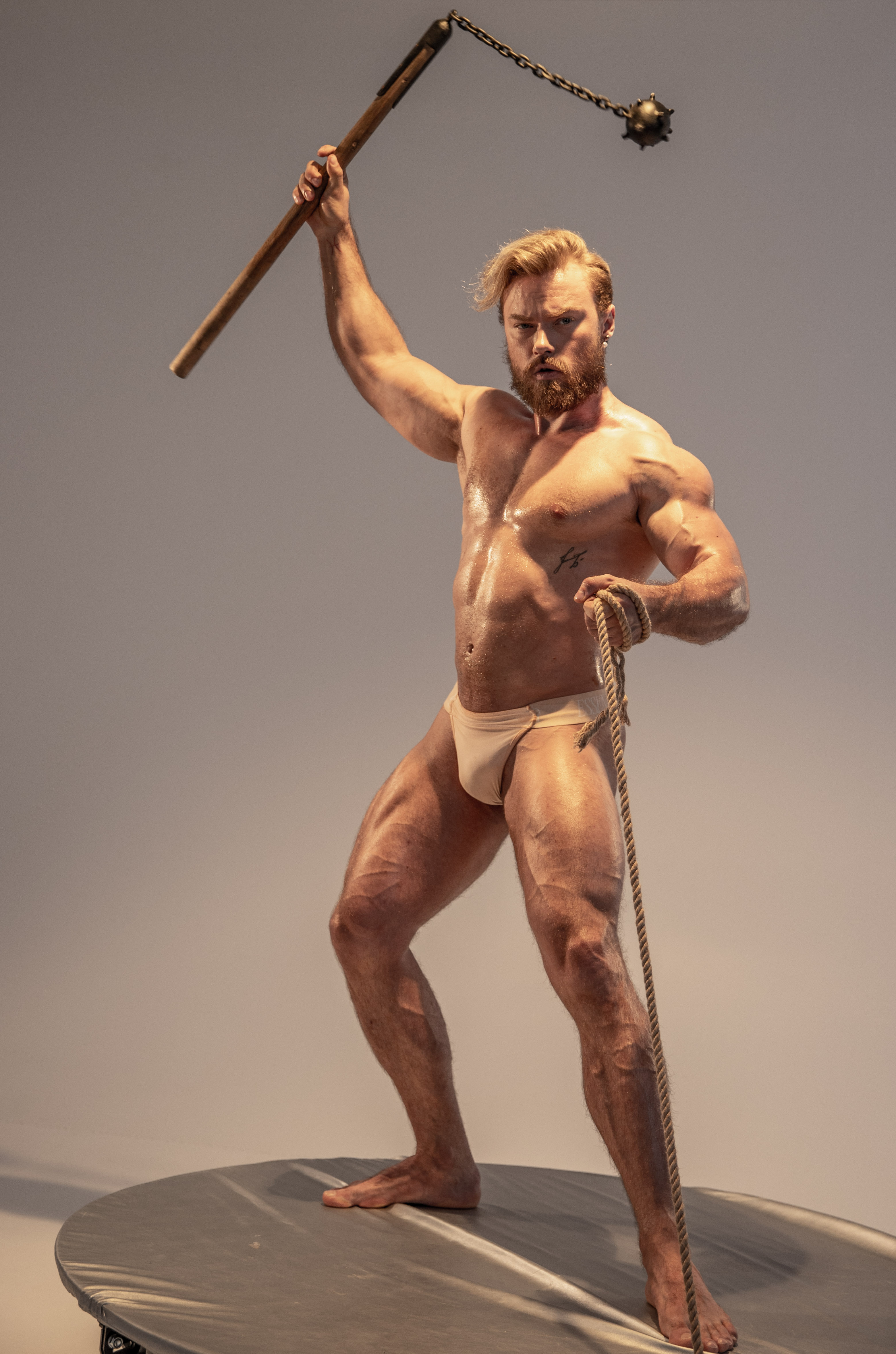 a muscular, bearded man stands on a small round platform in a studio setting. he is holding a flail weapon above his head with his right hand, the rope of which trails down to the platform beside his left foot. he is wearing only white underwear and has a tattoo on his left hip. the lighting is soft and diffused, highlighting his physique. his pose and expression convey a sense of strength and power.