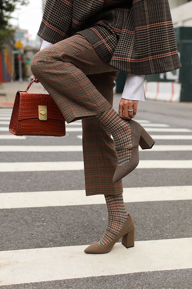 a stylish woman in a brown and beige plaid pantsuit walks across a crosswalk. she is wearing matching patterned socks and heels and carrying a crocodile handbag. the image is a street style shot that captures the woman's sophisticated autumnal outfit.