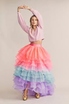 a woman in a pink sweater and a multicolored tulle skirt poses with her arms up in the air. the skirt is very full and layered, with shades of pink, purple, and blue. her blonde hair is styled in a loose wave. the woman's pose is playful and whimsical, and her outfit is both stylish and feminine.