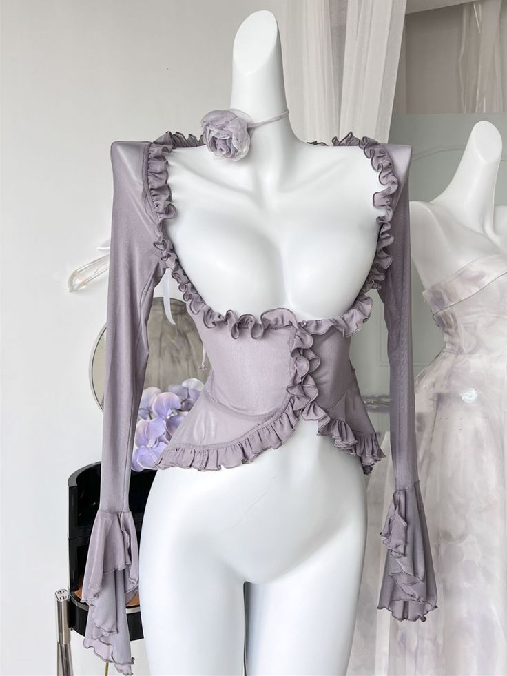 a purple corset top with long sheer sleeves and a ruffled neckline is displayed on a white mannequin in a studio setting. the top has a sweetheart neckline and a pointed hemline. the mannequin is positioned in front of a white wall, and a white dress is partially visible in the background. the lighting is soft and diffused, creating a romantic and elegant atmosphere.