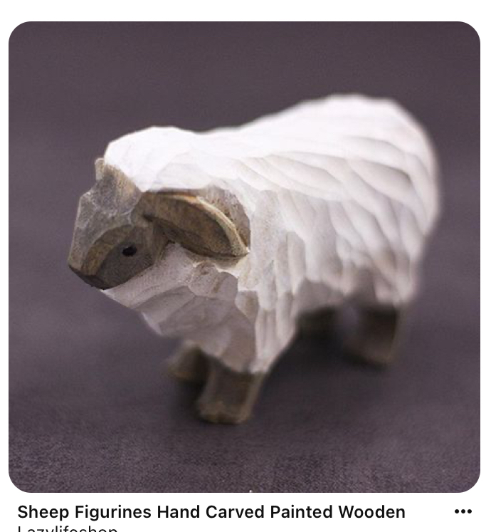 the image shows a handcarved and painted wooden sheep figurine. the sheep is white with a gray face and legs, and is standing on a dark gray surface. it is facing to the left of the image. the figurine is very detailed, with individual strands of wool carved into its body. the lighting is soft, creating a warm and inviting atmosphere.