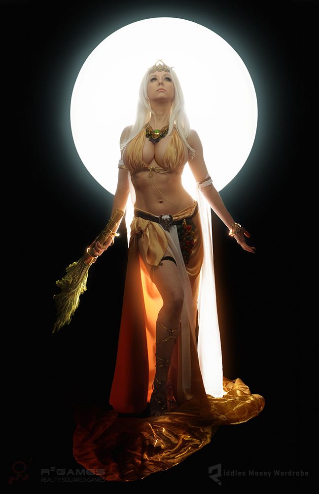 a woman in a golden goddess costume stands in front of a bright white circle of light, against a black backdrop. she holds a golden staff in her left hand and wears a golden crown. the costume is flowing and revealing, with a long, sheer skirt and a bra top. the lighting is dramatic and highlights the woman's curves. her expression is serene and powerful.